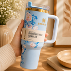 A hand holds a light blue Stanley Quencher H2.0 tumbler with floral graphics, a gold logo, and a white handle in a warm, styled indoor setting with a wooden table, dried flowers, and a framed print.