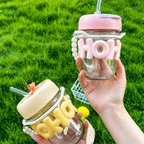 Two glass jars with pink and yellow letter decorations held by a hand against a grass background.