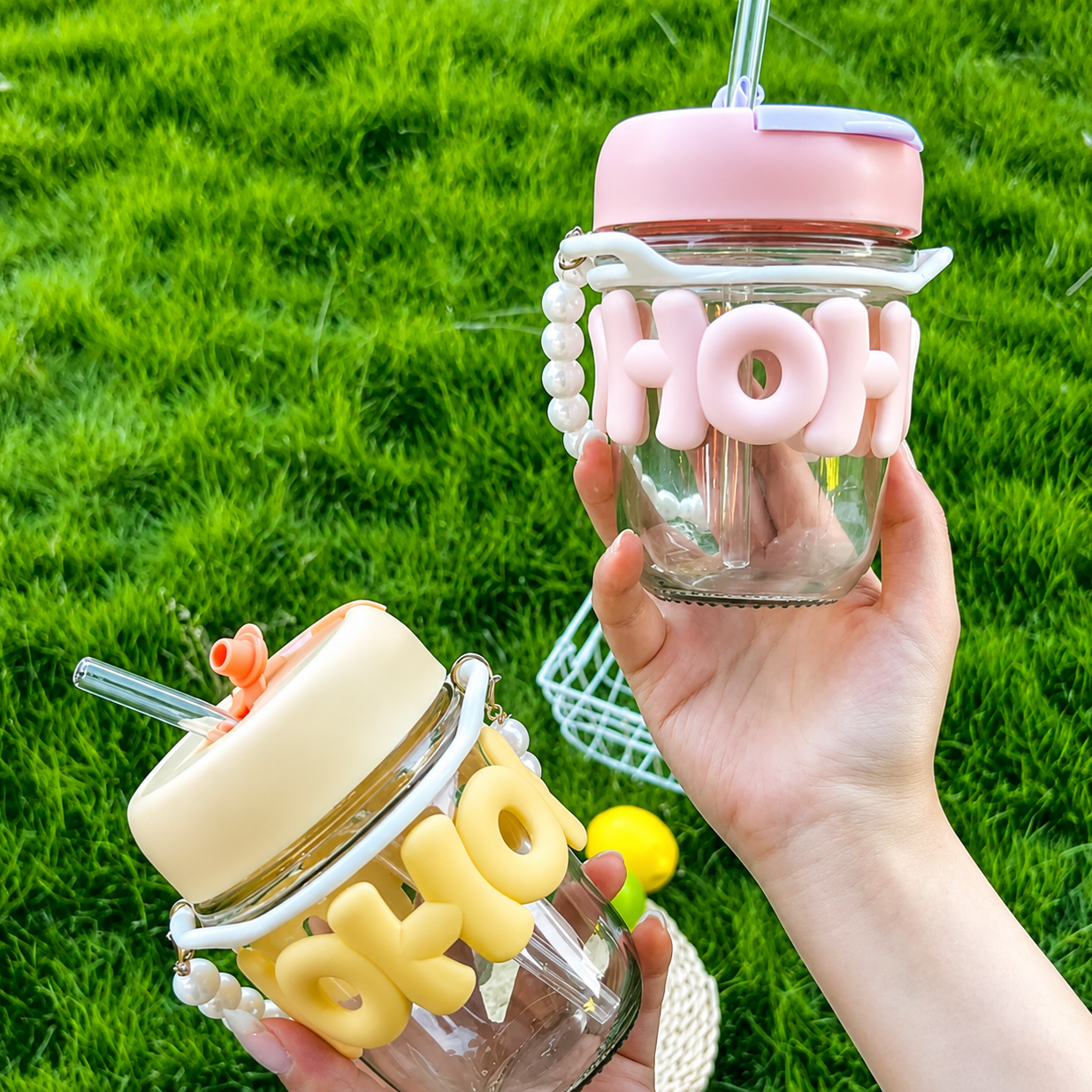Two glass jars with pink and yellow letter decorations held by a hand against a grass background.