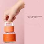 Hand holding an orange lock-shaped container against a pink background with text 'Life is not only colorful but also lovely'.