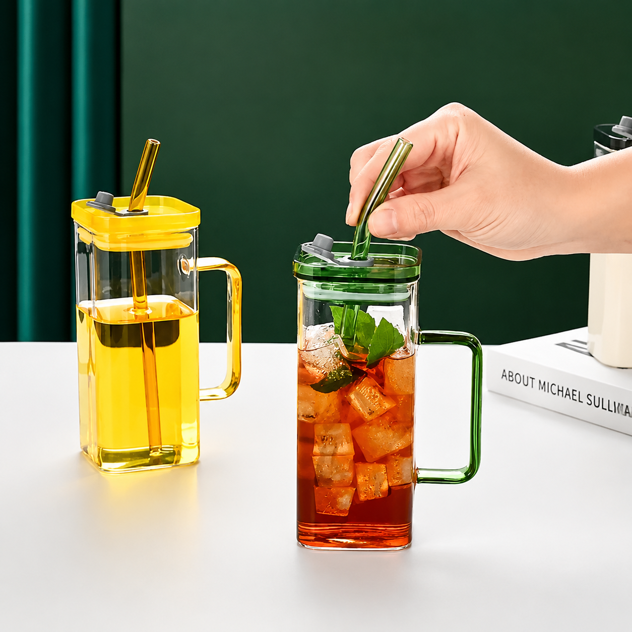 Person stirring a glass of iced tea with a green straw next to a yellow glass pitcher on a white surface.