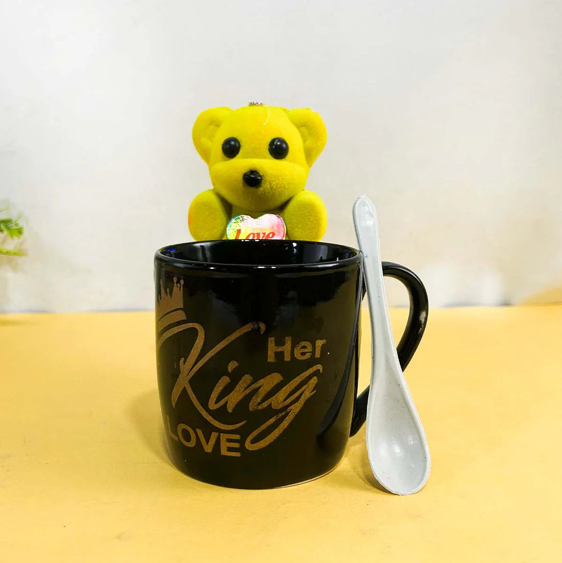 Black mug with 'Her King Love' text, , and spoon on a yellow surface.